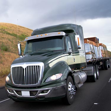 black semi-truck on road with green rolling hills and cloudy skies