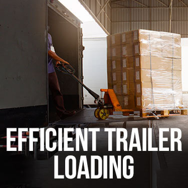 Warehouse worker pulling a pallet jack loaded with a fully stacked pallet of shrink wrapped boxes into a trailer.